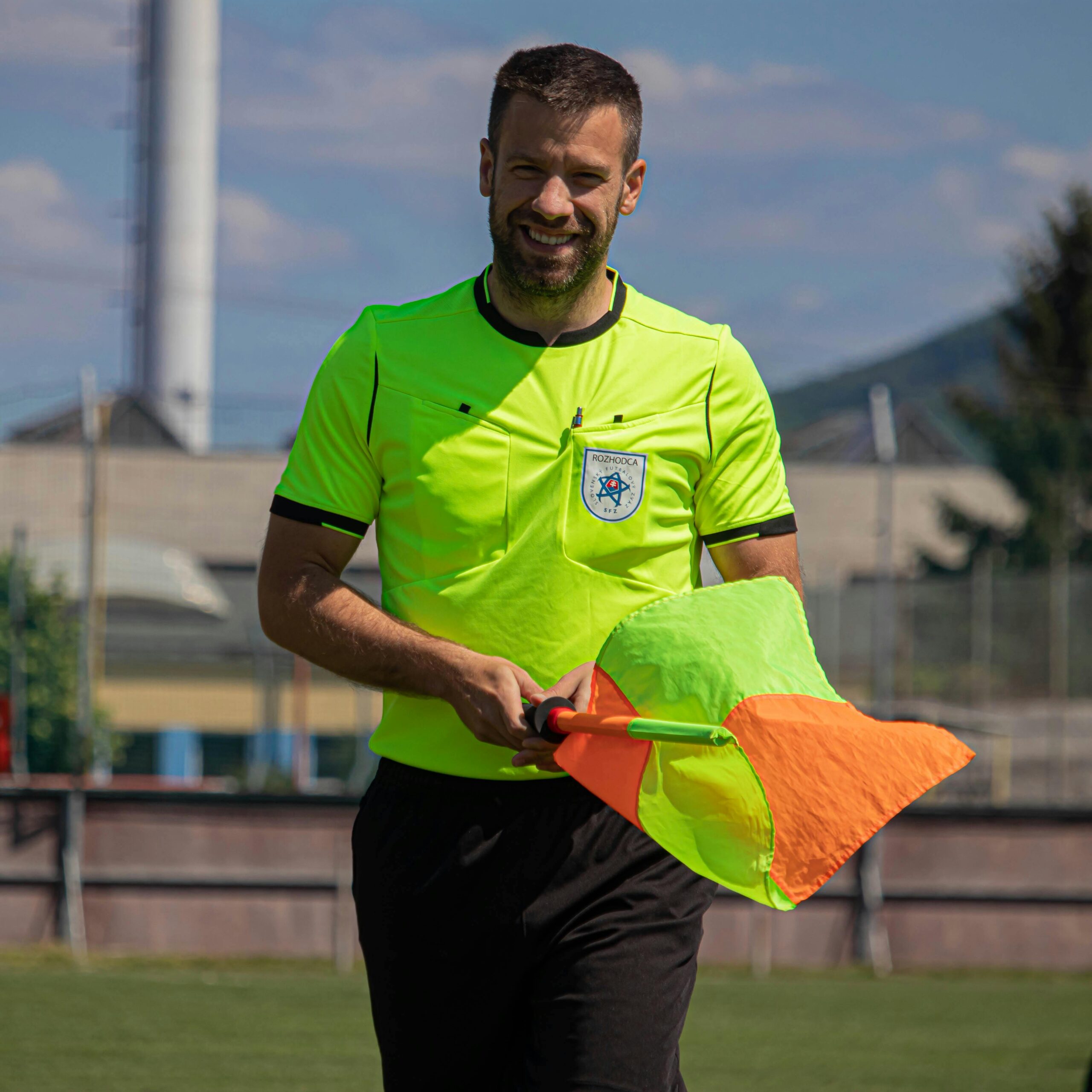 A smiling soccer referee holding a flag on a sunny day in Slovakia.