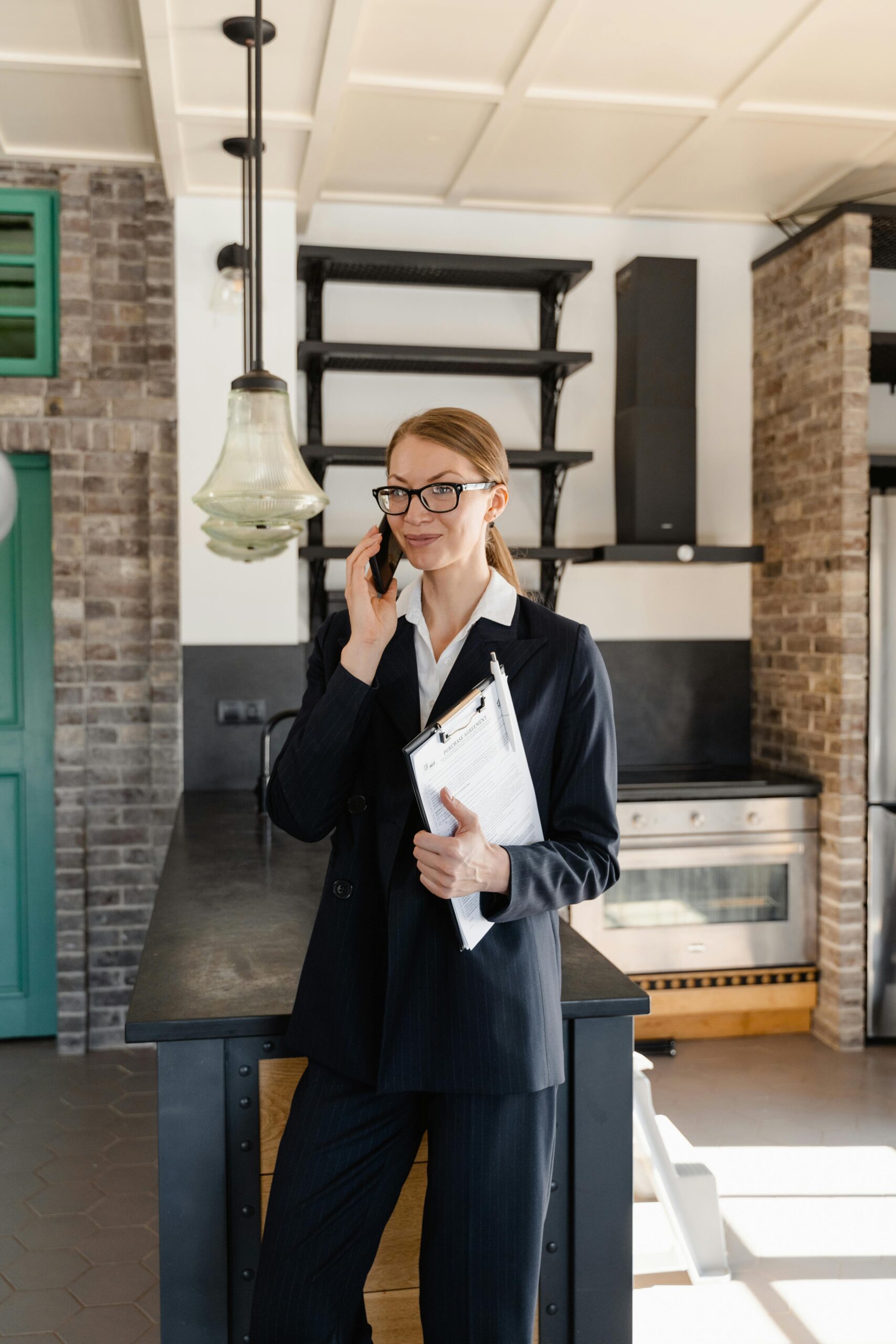 Confident female realtor in business attire on a call, smiling in a contemporary kitchen interior.